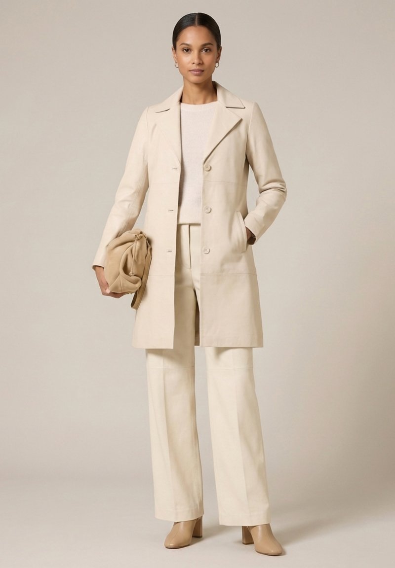 Femme portant un long manteau beige, un pantalon ample, un pull léger et des bottines beige, tenant un sac à main beige plié sur un fond beige.