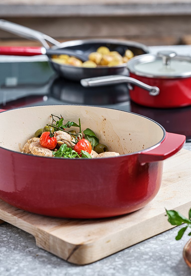 Olla de hierro fundido esmaltada en rojo con un interior blanco, llena de pollo, tomates cherry y verduras, sobre una tabla de cortar de madera.