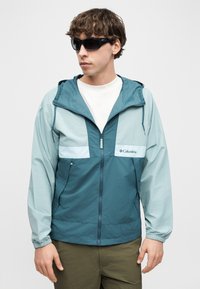 Νεαρός άνδρας που φοράει μαύρα γυαλιά ηλίου, ένα μπλε και τιρκουάζ μπουφάν Columbia με φερμουάρ και κουκούλα, και ελιάς χακί παντελόνια, κοιτάζοντας αριστερά μπροστά σε απλό φόντο.