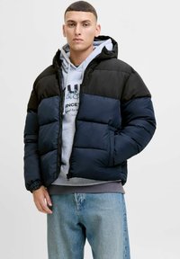 Puffjakke i sort og marineblå, med hætte, lynlås, quiltet tekstur og elastiske manchetter, lagd over en grå hoodie.