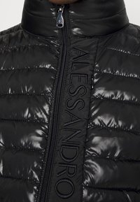 Musta toppatakki, jossa on korkea kaulus, tikattu pinta, vetoketjusuljenta ja brodeerattu logo edessä. Sileä, kiiltävä materiaali.