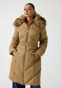 Cappotto imbottito lungo color sabbia con cappuccio in pelliccia sintetica, cuciture diagonali, vita con cintura e tasche laterali. Chiusura frontale con zip.