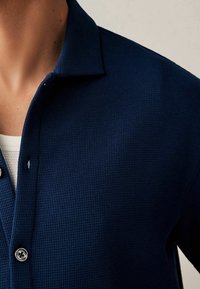 Chemise bleu marine à col en tissu texturé, avec une fermeture boutonnée sur le devant comportant deux boutons visibles. Motif côtelé subtil.