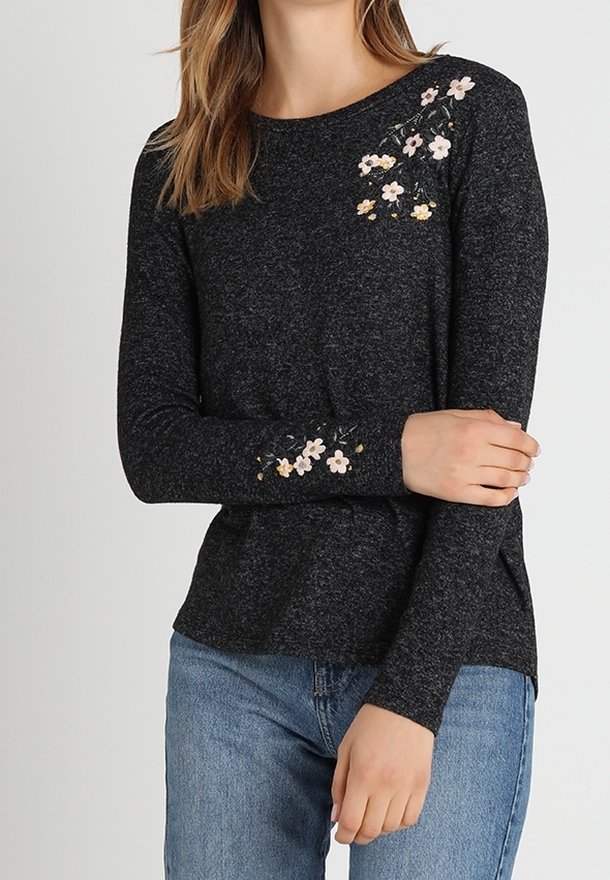 Femme portant un haut gris foncé à manches longues avec une broderie florale rose et jaune sur l'épaule et la manche, associé à un jean bleu.