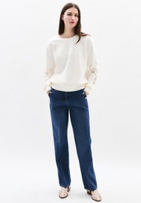 Felpa crema con dettagli in eyelet sulle maniche, abbinata a jeans a gamba larga blu scuro e sandali beige con lacci. Outfit semplice e casual.
