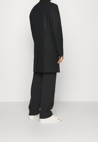 Zwarte wollen jas met een getailleerde pasvorm, hoge kraag en rugsplit, gecombineerd met losse zwarte broeken en witte sneakers met subtiele details.