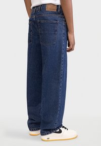 Φαρδιά τζιν σε σκούρο μπλε χρώμα, με δύο πίσω τσέπες και δερμάτινο ταμπελάκι στη μέση. Συνδυασμένο με λευκά αθλητικά παπούτσια.