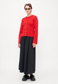 Cardigan rouge en tricot avec des manches longues, fermeture à boutons et poche. Associé à une jupe noire volumineuse et à des chaussures noires massives.