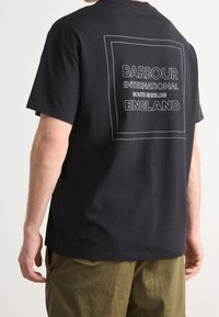 Černé bavlněné tričko s obdélníkovou bílou grafikou na zádech, na které je tučným písmem napsáno "Barbour International South Shields England".