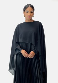 Czarny zestaw z satynową peleryną i plisowaną spódnicą. Materiał ma gładką teksturę i płynny krój. Złote kolczyki w kształcie kółek.