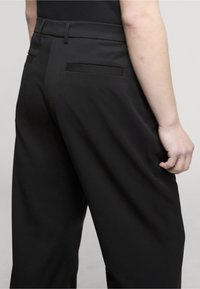 Person wearing loose black trousers and a black top, shown from the waist down with one hand relaxed by their side.