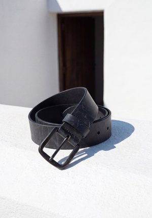 Un cinturón de cuero negro con una hebilla metálica rectangular está enrollado sobre una mesa blanca bajo una intensa luz solar.