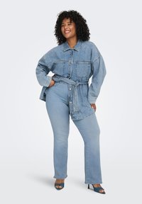 Γυναίκα με σγουρά μαλλιά που χαμογελά, φοράει oversized τζιν μπουφάν με ζώνη, τζιν παντελόνι ανοιχτού χρώματος και τυρκουάζ πέδιλα ανοιχτού δαχτύλου, στέκεται μπροστά σε λευκό φόντο.