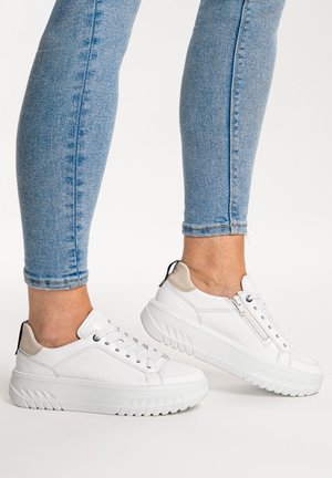 Ankle-length blue jeans worn with white platform sneakers featuring laces, side zipper, and beige heel accents on a neutral background.
