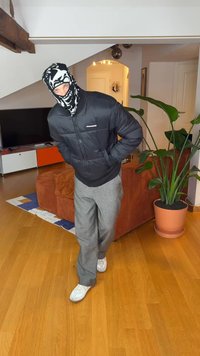 Black puffer jacket with zipper, grey wide-leg trousers, and white sneakers. Black and white skull-patterned mask. Indoor setting with plants.