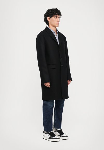Black long coat with three buttons, notch lapels, and side pockets. Worn over a light shirt, paired with blue jeans and black and white sneakers.