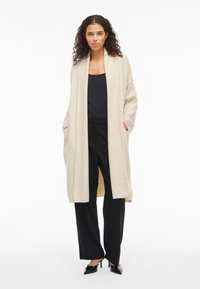 Γυναίκα με σγουρά μαλλιά, ντυμένη με μακρύ μπεζ κάρντιγκαν, μαύρο τοπάκι, μαύρο παντελόνι φαρδύ και μαύρα ψηλοτάκουνα παπούτσια με μυτερές μύτες, στέκεται με τα χέρια στις τσέπες.