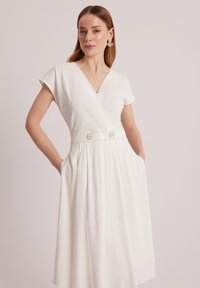 Biała sukienka z dekoltem w kształcie litery V, krótkimi rękawami, szwem w talii i dużymi guzikiem. Materiał miękko się układa, a po bokach znajdują się kieszenie, co zwiększa jej funkcjonalność.