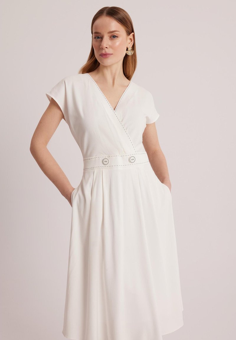 Biała sukienka z dekoltem w kształcie litery V, krótkimi rękawami, szwem w talii i dużymi guzikiem. Materiał miękko się układa, a po bokach znajdują się kieszenie, co zwiększa jej funkcjonalność.