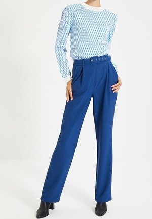 Mannequin portant un pantalon bleu taille haute avec ceinture, un haut à manches longues à motifs et des bottines noires à talons sur fond blanc.
