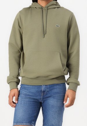Homme portant un sweat à capuche vert olive avec poche avant et logo Lacoste, associé à un short en jean bleu.