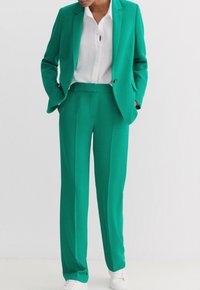 Completo due pezzi color turchese realizzato in tessuto morbido, composto da un blazer su misura e pantaloni dritti, abbinato a una camicia bianca con colletto e scarpe da ginnastica bianche.