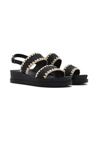 Sandales tressées noires avec deux larges brides, accents beige en forme de vagues, sangle de cheville ajustable et une épaisse semelle noire. Détails en matériau texturé visibles.