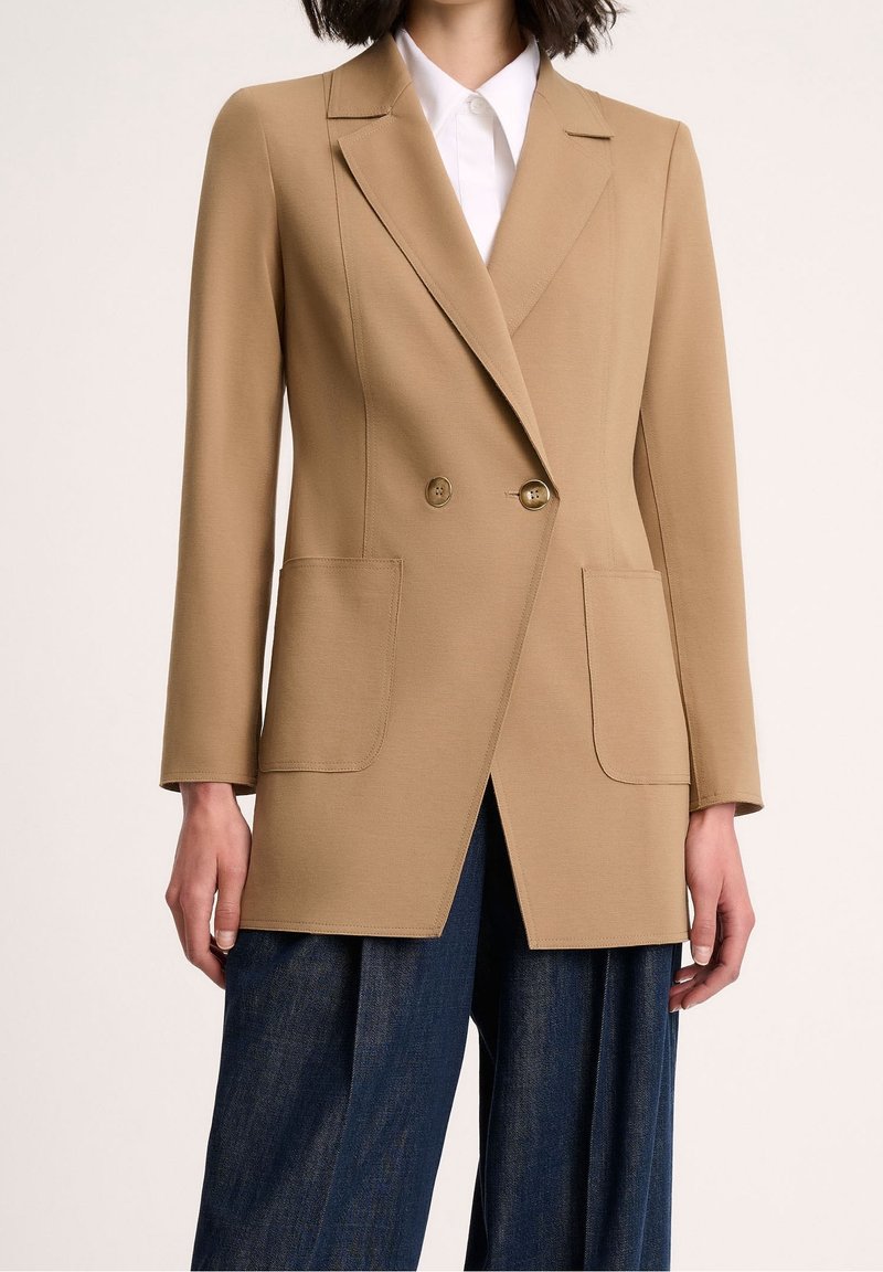 Beiger zweireihiger Blazer mit zwei Fronttaschen, eingekerbten Revers und goldenen Knöpfen. Der Stoff hat eine glatte, strukturierte Textur.