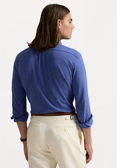 Camisa azul de manga longa com punhos dobrados, tecido leve e corte descontraído, combinada com calças de cor creme com bolsos traseiros.