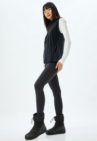 Mustaan fleece-liivi, jossa on huppu, yhdistettynä mustiin legginsseihin ja mustiin eristäviin saappaisiin. Asusteessa on sileät pinnat ja tiukka muotoilu.