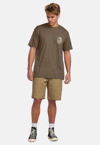 Brown cotton t-shirt with a small graphic on the chest, paired with beige shorts and black high-top sneakers with white laces.