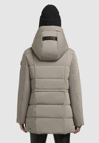Beige pufferjakke med hætte, der har kviltet tekstur og et logo patch. Accepteret af en sort strikket beanie. Vist set fra ryggen.