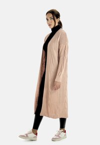 Lång, öppen cardigan i ljusrosa med kabelstickad textur, parat med en svart polotröja och figurnära svarta leggings, samt flerfärgade sneakers.