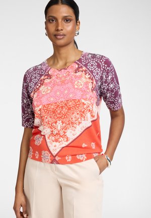 Vrouw draagt een korte mouwen top met patroon in oranje, roze en paars met crèmekleurige broek, zilveren oorbellen en een zilveren armband.