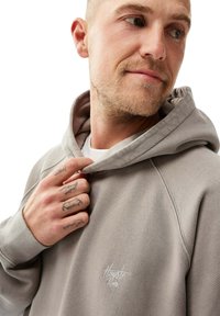 Ljusgrå hoodie med en känguruficka, ribbade muddar och en broderad logotyp på bröstet. Tyget verkar mjukt med en avslappnad passform.