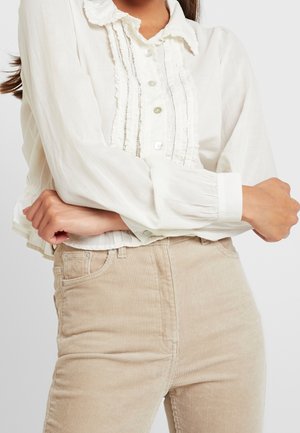 Femme portant un chemisier blanc à manches longues avec volants et boutons, associé à un pantalon beige en velours côtelé taille haute, les bras croisés.