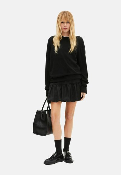 Zwart oversized trui met lange mouwen, gecombineerd met een zwart rokje met ruches. Accessoires omvatten een gestructureerde zwarte handtas en zwarte sokken met schoenen.