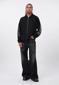 Čierna mikina na zips s textúrovaným sieťovaným detailom na rukávoch, v kombinácii s tmavými džínsami širokých nohavíc a čiernymi platformovými topánkami. Jednoduchý, na bežné nosenie určený dizajn.