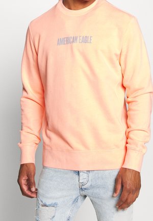 Mężczyzna ubrany w łososiową bluzę z długim rękawem z napisem "AMERICAN EAGLE" oraz jasnoniebieskie podarte dżinsy, stojący na tle białego tła.