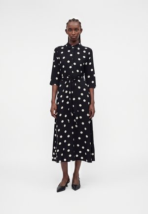 Femme debout portant une robe midi noire à pois avec des boutons, des manches trois-quarts, une ceinture à la taille et des talons noirs pointus.