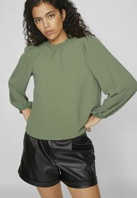 Groene, gestructureerde blouse met opgeblazen mouwen, gatherede hals en elastische manchetten, gecombineerd met zwarte nepleerenshort. Eenvoudig en hedendaags ontwerp.