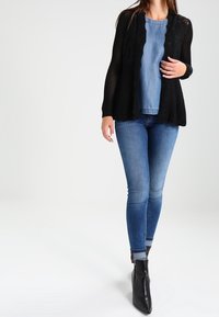 Svart öppenstickad cardigan över en blå denimtop, kombinerat med smala blå jeans och svarta ankelstövlar. Har en avslappnad passform och långa ärmar.