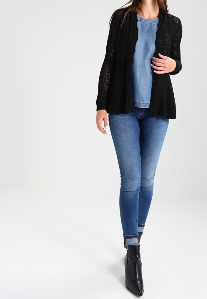 Svart öppenstickad cardigan över en blå denimtop, kombinerat med smala blå jeans och svarta ankelstövlar. Har en avslappnad passform och långa ärmar.