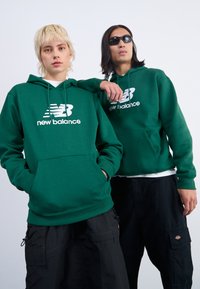 Sweatshirts à capuche verts avec le logo blanc "New Balance". L'un est associé à un pantalon cargo noir ; l'autre avec un t-shirt noir et des lunettes de soleil.