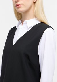 Zwarte mouwloze jurk met een V-hals, gelaagd over een witte blouse met kraag. Gladde stof zonder zichtbare versieringen of patronen.