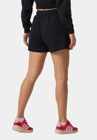 Person trägt schwarze Shorts mit elastischem Bund, schwarzen kurz geschnittenen Pullover und rote Adidas-Sneaker mit weißen Socken, steht mit dem Rücken zur Kamera.