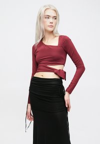 Bordo dugi rukav uski top s kvadratnim izrezom, vezan na struku, u kombinaciji s crnom suknjom s nabranim detaljima i vezicama.