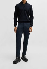 Mörk marinblå zip-tröja med långa ärmar, matchad med slim-fit byxor i en mörkare nyans, och robusta svarta ankelstövlar.