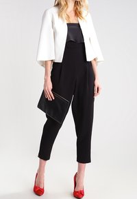 Vrouw in een zwarte jumpsuit, met een witte cropped jack, rode hakken en een zwarte clutch, tegen een effen lichte achtergrond.