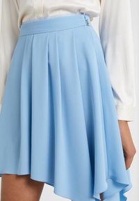 Falda asimétrica de color azul claro, hecha de tela fluida. Presenta pliegues y un cierre con cremallera lateral. Textura suave con un acabado liso.